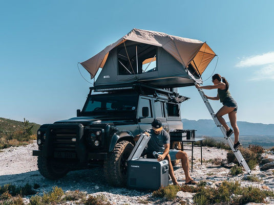 Woman setting up Front Runner Roof Top Tent on a rugged vehicle while man unpacks camping gear in a scenic outdoor landscape