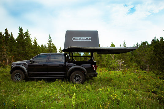 Black pickup truck equipped with Freespirit Recreation Odyssey Series Black Top Hard Shell Rooftop Tent parked in a forest clearing.