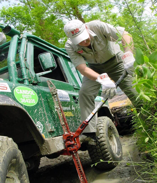Man using Hi-Lift Jack 60 Cast/Steel to lift a muddy green off-road vehicle outdoors.