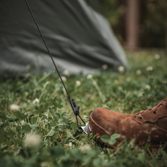 Close-up view of a Gazelle Tents T4 Hub Tent's ground stake with tension rope and a hiker's boot in the foreground, camping equipment on grass.