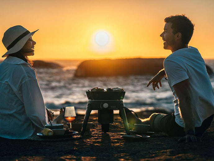 Couple enjoying sunset at beach with Front Runner Safari Chef 30 HP Portable 5 Piece Gas Barbeque Camp Cooker by CADAC.
