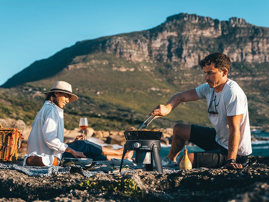 Couple enjoying outdoor cooking with Front Runner Safari Chef 30 HP Portable Gas Barbeque by CADAC during a camping trip.