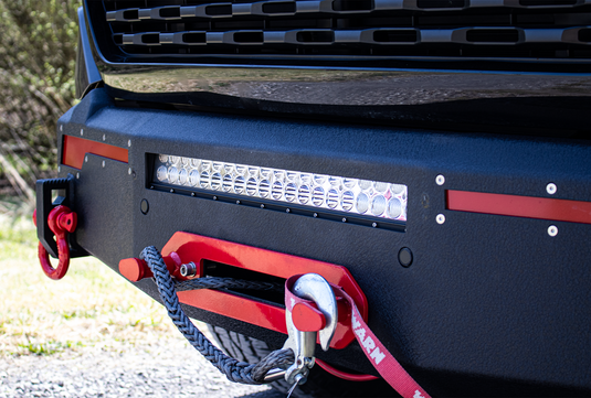 Alt text: "Close-up of a Fishbone Offroad 2019-2023 Ram Pike Front Bumper featuring an integrated LED light bar and red tow hooks on a black truck."