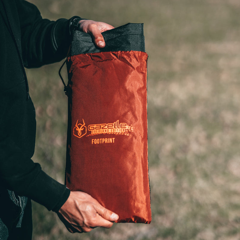 Load image into Gallery viewer, Alt text: &quot;Person holding a folded Gazelle Tents T4 Tent Footprint in a red storage bag with the brand logo clearly visible.&quot;
