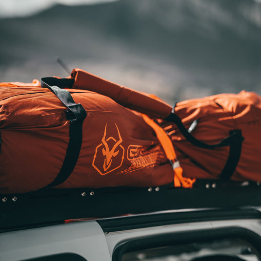 Alt-text: "Gazelle Tents T4 orange water-resistant duffle bags on a vehicle roof rack with mountainous background."