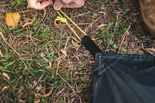 Person securing a corner of a Gazelle Tents T4 Plus & T8 Footprint to the ground with a yellow stake on grassy terrain.