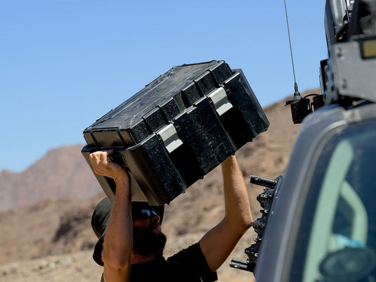 Man loading Front Runner Wolf Pack Pro Hi-Lid storage box onto a vehicle in a desert terrain.