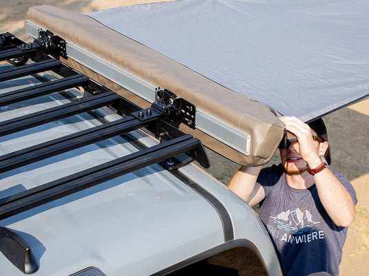 Person installing a Front Runner Slimsport Side Mount Accessory Bracket on a vehicle roof rack system underneath an extended awning.