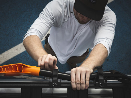 Person securing an orange spade with a Front Runner Ratcheting Spade/Shovel Mount to a black roof rack