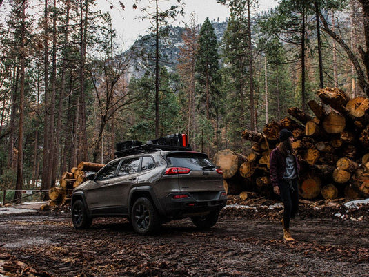 Jeep Cherokee KL equipped with Slimline II Roof Rail Rack Kit by Front Runner parked in a forest near a pile of logs with a person standing alongside.