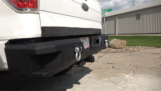 Alt text: "Fishbone Offroad rear step bumper installed on a 2009-2014 white Ford F-150, featuring tow hooks and a textured black finish."