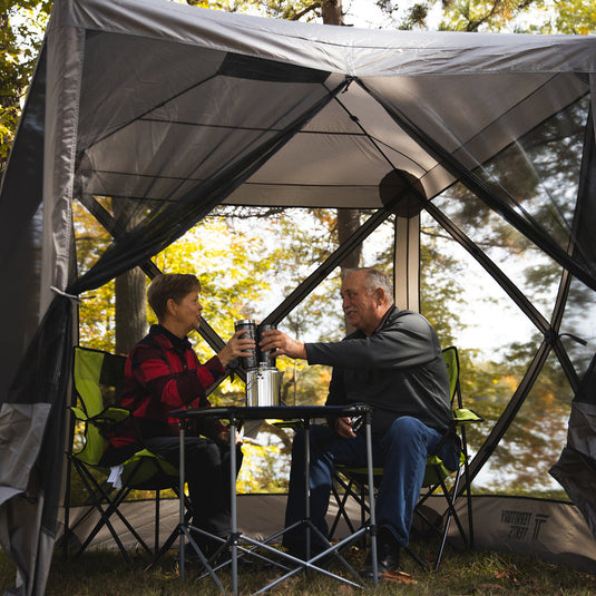 Alt text: Two people sitting under a Territory Tents 4-Sided Portable Screen Tent enjoying beverages in a forest campsite.