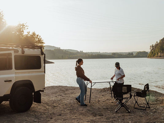Alt text: "Two people setting up a Front Runner Under Rack Table next to a white off-road vehicle by the lakeside during a camping trip."