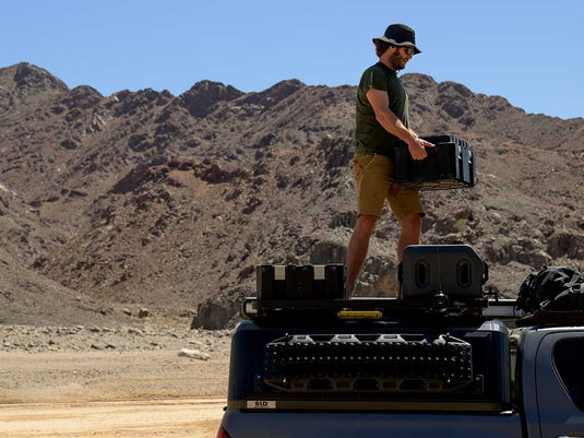 Man holding a Front Runner Wolf Pack Pro Hi-Lid storage box on top of off-road vehicle with mountainous desert in the background.