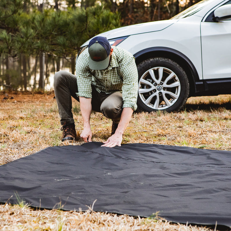 Load image into Gallery viewer, Man setting up a black Gazelle T3X Overland Edition Tent footprint near a vehicle in a campsite setting.
