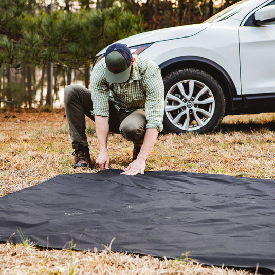 Man setting up a black Gazelle T3X Overland Edition Tent footprint near a vehicle in a campsite setting.