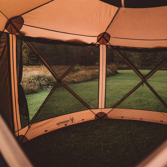 Gazelle Tents G6 Deluxe 6-Sided Portable Gazebo set up in a grassy field with a view of trees and nature through its mesh windows.