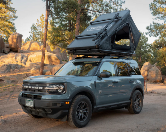 Alt text: "Freespirit Recreation Odyssey/Evolution rooftop tent on a SUV with cross bar kits, parked outdoors with pine trees and rocks in the background."