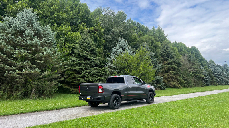 Load image into Gallery viewer, Alt text: &quot;2019-2023 Ram 1500 equipped with Fishbone Offroad Pike Rear Bumper parked on a gravel road beside a forested area.&quot;
