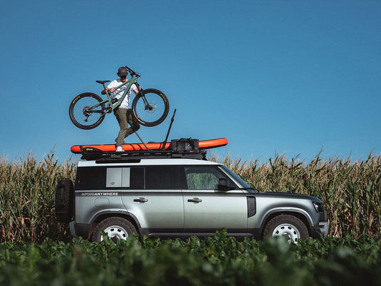 Cyclist mounting a mountain bike onto the Front Runner Pro Bike Carrier on a vehicle roof, parked in a field.