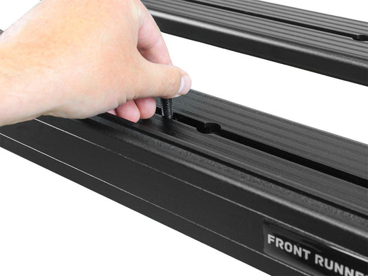 Installation of a Front Runner Slimline II Roof Rail Rack on a Jeep Grand Cherokee, showing hand tightening a bolt on the sturdy black rack rails engraved with the Front Runner logo.