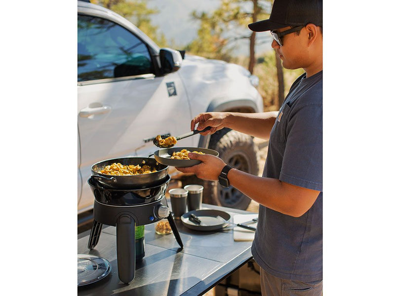 Load image into Gallery viewer, Man using Front Runner Safari Chef 30 HP portable gas barbeque by CADAC to cook food outdoors next to a vehicle on a camping trip.
