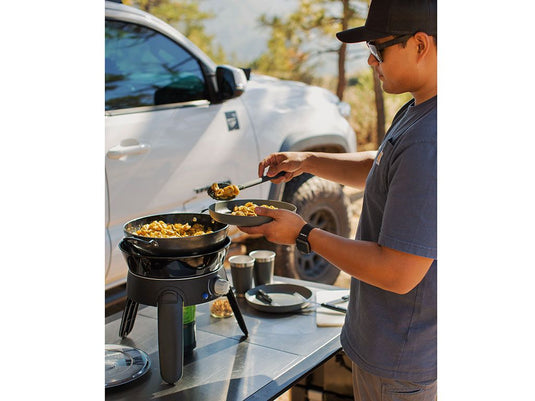 Man using Front Runner Safari Chef 30 HP portable gas barbeque by CADAC to cook food outdoors next to a vehicle on a camping trip.