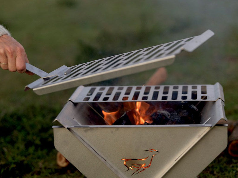 Load image into Gallery viewer, Front Runner BBQ/Fire Pit being used outdoors with a person lifting the grill top to reveal glowing embers
