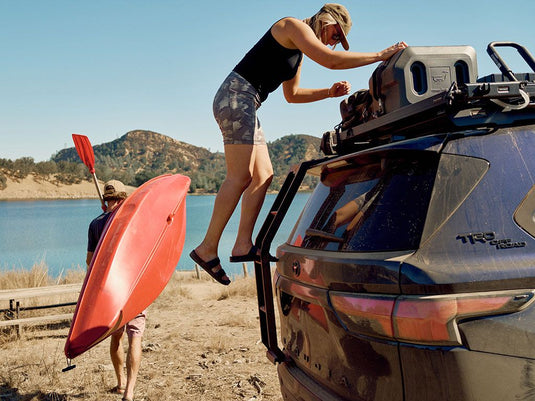 Person climbing Front Runner ladder on a Toyota Sequoia by a lake with another person carrying a kayak