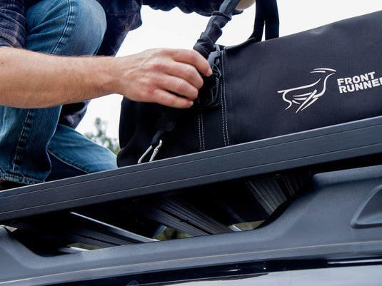 Person securing a black Front Runner Stratchit to a vehicle's roof rack for safe transportation of cargo.