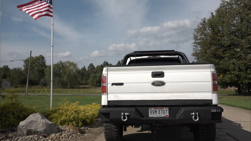 Load image into Gallery viewer, Fishbone Offroad rear step bumper on a 2009-2014 Ford F-150, showcasing durability and design with towing hooks and American flag in the background
