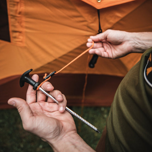 Close-up of hands setting up the Gazelle Tents T4 Hub Tent Overland Edition by securing tent peg with orange guyline.