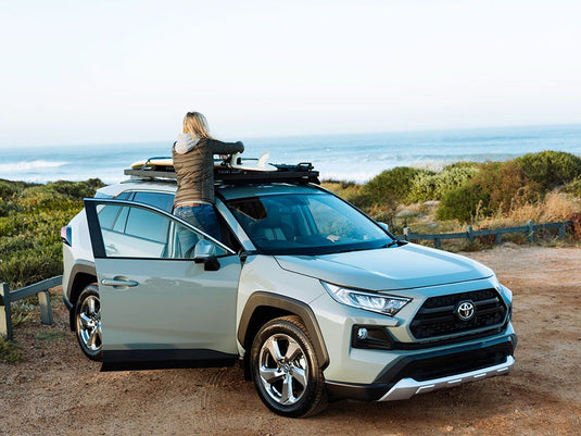 Woman standing next to a 2019 Toyota RAV4 equipped with Front Runner Slimline II Roof Rack Kit, parked near the coastline.