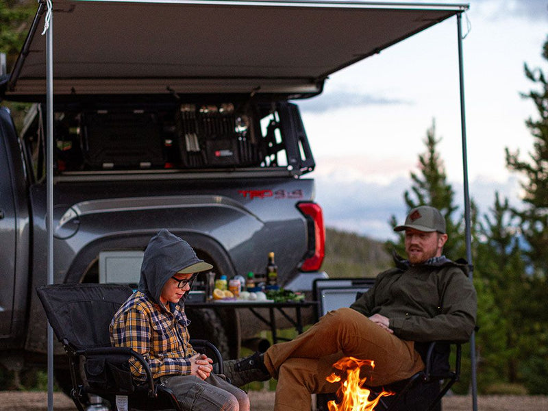 Load image into Gallery viewer, Alt text: Two people relaxing by a campfire with a 2022 Toyota Tundra CrewMax featuring a Front Runner Pro Bed Rack Kit in the background.
