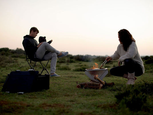 Two people relaxing by a portable Front Runner BBQ/Fire Pit during a twilight outdoor adventure.