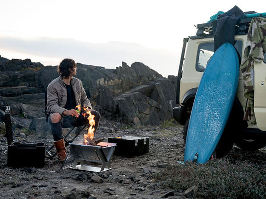 Man cooking on a portable Front Runner BBQ/Fire Pit near a vehicle with camping and surf gear outdoors.
