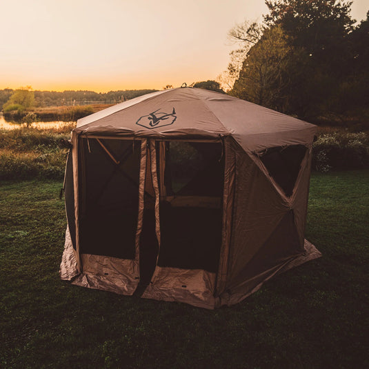 Gazelle Tents G6 Deluxe 6-Sided Portable Gazebo set up on grass at dusk, with surrounding trees and a serene lake in the background.