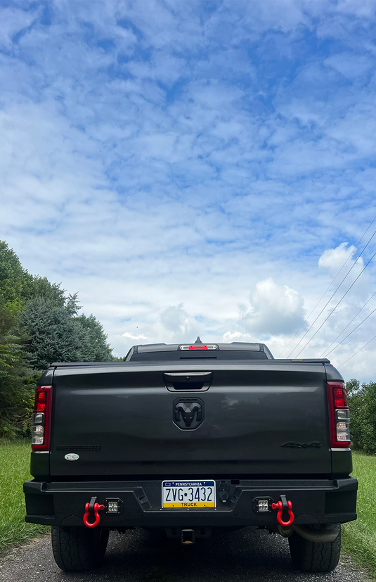 Alt text: "2019 Ram 1500 truck with Fishbone Offroad Pike Rear Bumper upgrade, equipped with tow hooks, seen from the back on a cloudy day."