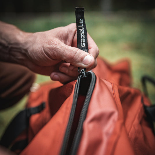 Close-up of a person's hand holding a Gazelle Tents T4 Water-Resistant Duffle Bag zipper with logo branding in an outdoor setting.
