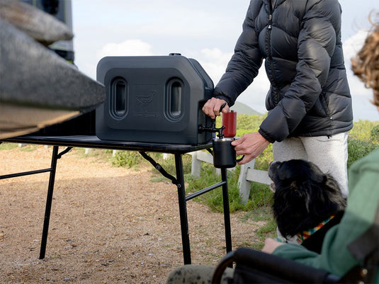 Alt text: "Person using Front Runner Pro 20L Water Tank with Mounting System to pour water into a cup, mounted beside a camping table outdoors, with a dog nearby."