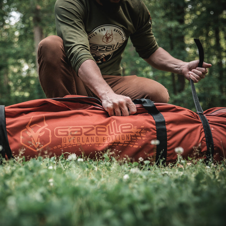 Load image into Gallery viewer, Alt text: &quot;Person in a forest setting securing a large orange Gazelle Tents T4 Overland Edition water-resistant duffle bag.&quot;
