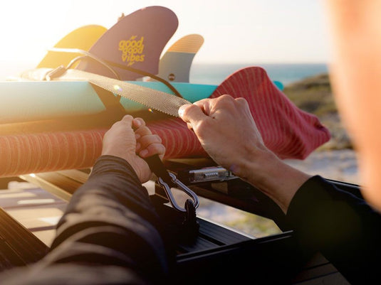 Person securing surfboards on car roof rack with Front Runner Black Tie Down Rings at sunset