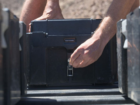 Alt text: "Close-up of hands securing a Front Runner Wolf Pack Pro storage box with MKII Rack Mounting Brackets on a vehicle's rack system."