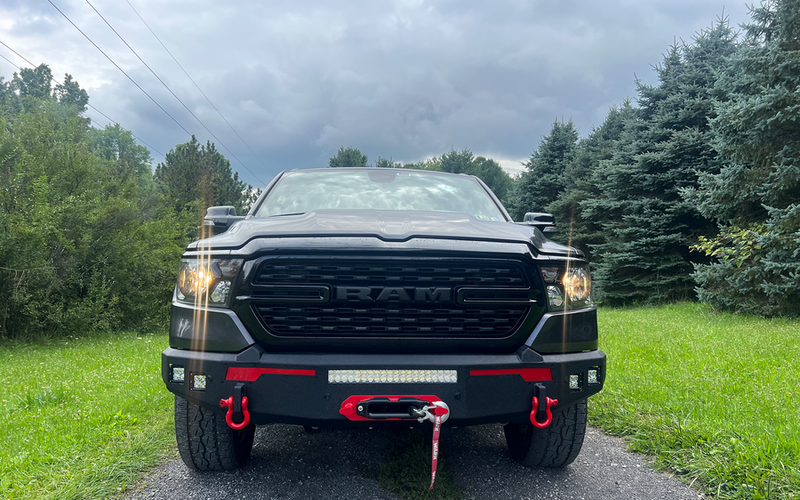 Load image into Gallery viewer, Alt text: &quot;Black 2019 Ram 1500 truck with Fishbone Offroad Pike Winch Plate, prominently displayed on front bumper with red tow hooks, parked outdoors.&quot;
