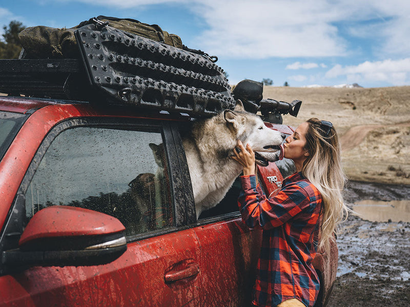 Load image into Gallery viewer, Front Runner Recovery Device and Gear Holding Side Brackets mounted on a red off-road vehicle with traction boards attached, under clear skies with a woman affectionately greeting a large dog.
