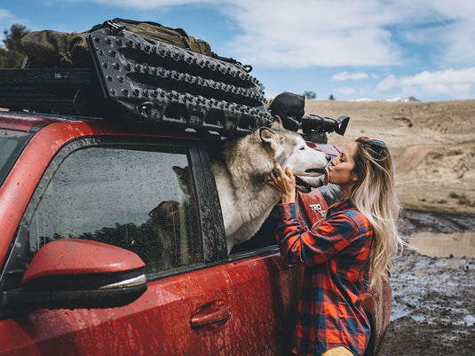 Front Runner Recovery Device and Gear Holding Side Brackets mounted on a red off-road vehicle with traction boards attached, under clear skies with a woman affectionately greeting a large dog.
