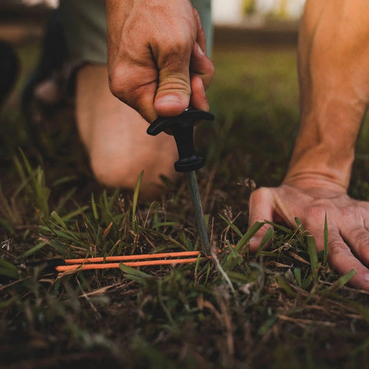 Person hammering a Gazelle Tents all-terrain stake into the ground with other stakes lying on the grass.