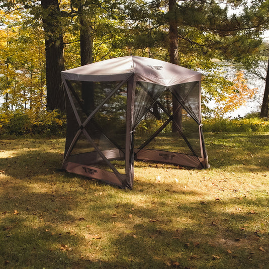 Alt text: "Territory Tents 4-Sided Portable Screen Tent set up on a grassy area with trees and a lake in the background, ideal for outdoor activities and camping."