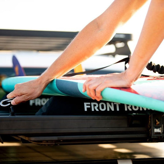 Person securing surfboard with Front Runner Rack Pad Set on vehicle roof rack