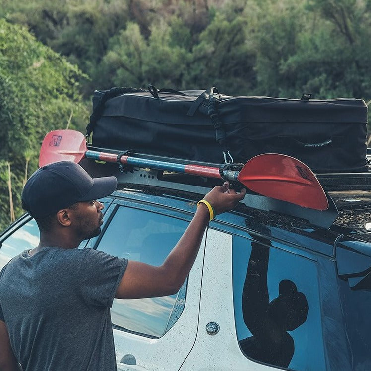 Load image into Gallery viewer, Man securing a red Front Runner Ratcheting Spade/Shovel Mount to a vehicle roof rack for off-road equipment storage
